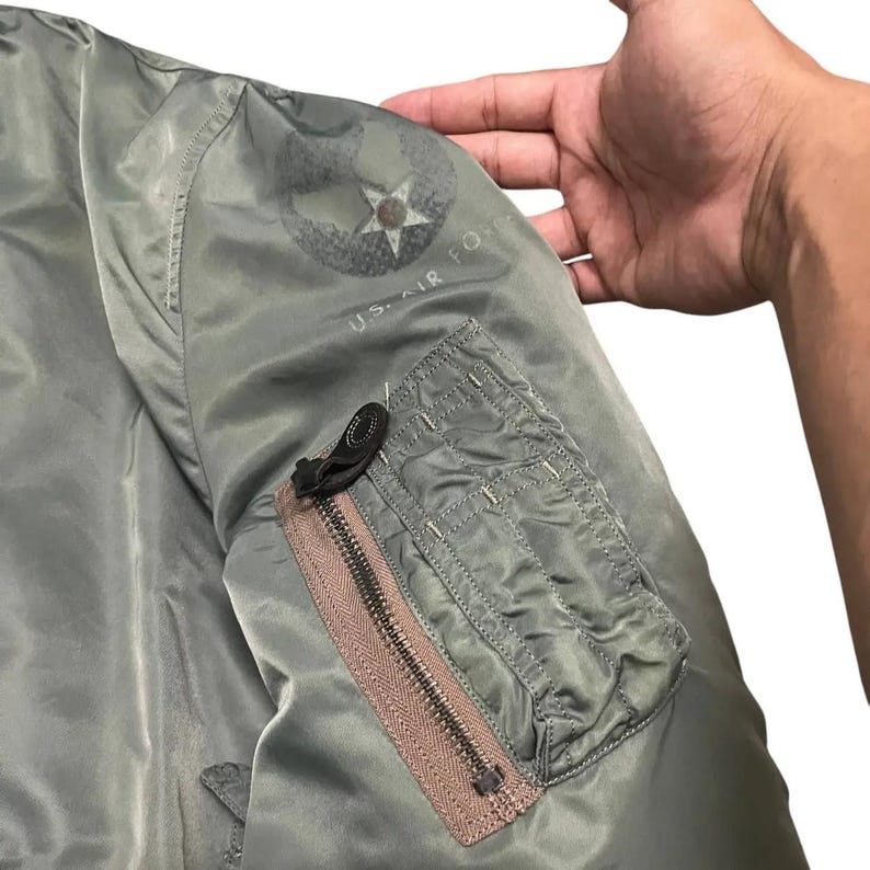 Puede incluir: Primer plano de una manga de chaqueta bomber verde oliva. Cuenta con un bolsillo con cremallera con una cremallera marr&oacute;n y una leng&uuml;eta negra. La chaqueta tiene un emblema de la Fuerza A&eacute;rea de EE. UU. en la manga superior.
