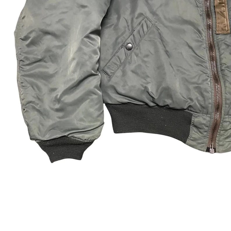 Puede incluir: Primer plano de una chaqueta bomber verde oliva. La chaqueta tiene pu&ntilde;os y dobladillo acanalados, una cremallera frontal y un bolsillo con cierre de bot&oacute;n a presi&oacute;n. Parche de cuero marr&oacute;n en la parte superior derecha.