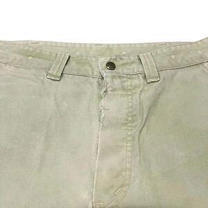 Op de afbeelding: Een lichtgroene broek met een knoopsluiting en een enkele knoop bovenaan de knoopsluiting.