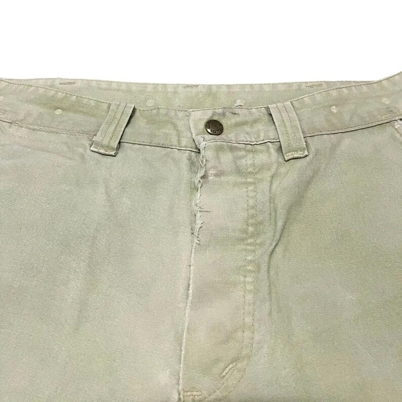 Op de afbeelding: Een lichtgroene broek met een knoopsluiting en een enkele knoop bovenaan de knoopsluiting.