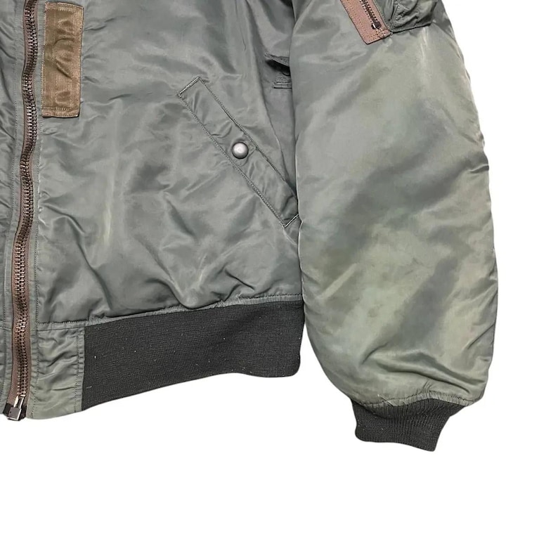 Puede incluir: Primer plano de una chaqueta bomber verde oliva. La chaqueta presenta un parche marr&oacute;n en la parte superior izquierda, un bolsillo con cremallera y un dobladillo y pu&ntilde;os de canal&eacute; verde oscuro. La chaqueta es de un material liso, posiblemente nailon.
