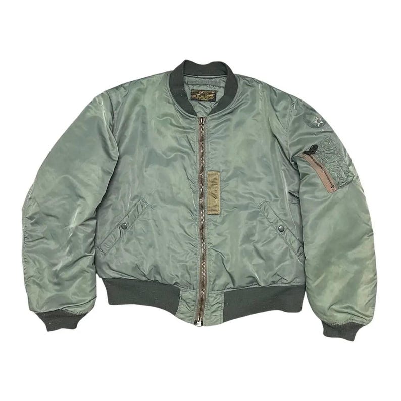 Puede incluir: Chaqueta bomber verde salvia con cuello, pu&ntilde;os y dobladillo de canal&eacute; verde oscuro. La chaqueta tiene cremallera frontal, dos bolsillos delanteros y un bolsillo peque&ntilde;o en la manga izquierda. Un emblema de estrella es visible.