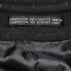 May include: Close-up of a black garment with a label. The label reads "COMPOSITION SUR ETIQUETTE ANNEXE, COMPOSITION: SEE ATTACHED LABEL". The fabric appears to be a dark, textured material.