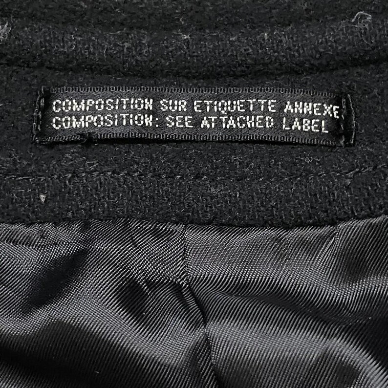 May include: Close-up of a black garment with a label. The label reads "COMPOSITION SUR ETIQUETTE ANNEXE, COMPOSITION: SEE ATTACHED LABEL". The fabric appears to be a dark, textured material.