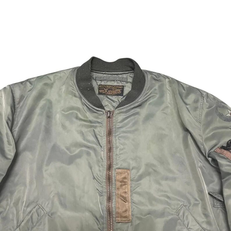 Puede incluir: Chaqueta bomber verde salvia con cuello y pu&ntilde;os de canal&eacute; verde oscuro. La chaqueta presenta cierre de cremallera completa, un parche marr&oacute;n y un peque&ntilde;o parche en la manga. La chaqueta est&aacute; hecha de un material liso y brillante.