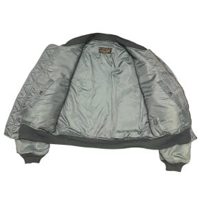 Puede incluir: Vista abierta de una chaqueta bomber verde salvia con un exterior acolchado y un interior liso, similar al sat&eacute;n. La chaqueta tiene cuello, pu&ntilde;os y dobladillo de canal&eacute; negro. En el interior se ve una peque&ntilde;a etiqueta.