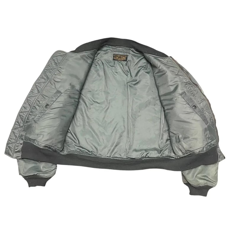 Puede incluir: Vista abierta de una chaqueta bomber verde salvia con un exterior acolchado y un interior liso, similar al sat&eacute;n. La chaqueta tiene cuello, pu&ntilde;os y dobladillo de canal&eacute; negro. En el interior se ve una peque&ntilde;a etiqueta.