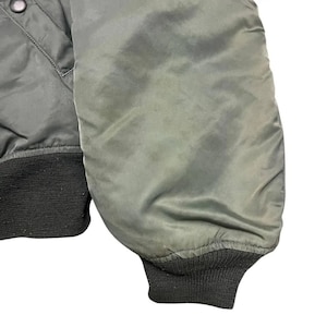 Puede incluir: Primer plano de una manga de chaqueta bomber verde. La manga est&aacute; hecha de un material liso y ligeramente brillante. El pu&ntilde;o es de punto acanalado verde oscuro. La chaqueta parece ser vintage o de estilo militar.