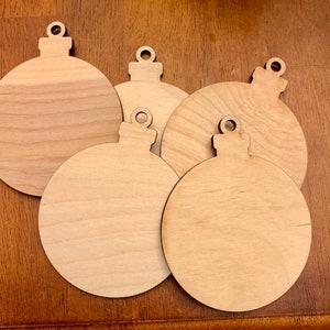 May include: A collection of five unfinished wooden Christmas ornament cutouts. Each ornament is round with a bulb shape and a small loop at the top for hanging. The wood has a light, natural color and visible grain.
