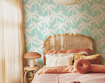 May include: A bedroom scene featuring a bed with pink bedding and pillows. The headboard is made of woven rattan. The wall is decorated with a teal wallpaper with a repeating pattern of pink and white palm trees. A small side table with a mushroom-shaped lamp is next to the bed.