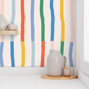 May include: A cream-colored wall with vertical stripes in various colors, including blue, green, yellow, and red. A gray pitcher and small cup sit on a wooden tray. A small shelf holds a cup.
