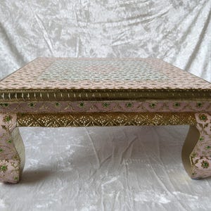 Può includere: Tavolino quadrato rosa e oro. Il piano presenta un motivo ripetuto di forme bianche e verdi. I bordi sono rifiniti in oro e le gambe curve sono decorate con motivi floreali. Il tavolo è alto circa 30 cm.