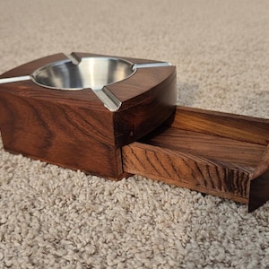 May include: A wooden ashtray with a stainless steel bowl and a pull-out drawer. The ashtray is made of dark brown wood and has a curved design. The drawer has a small brass knob. The ashtray is designed for cigars or cigarettes.