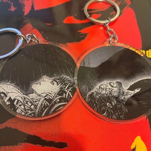 May include: Two round keychains with black and white illustrations of people. One keychain features a person lying down with their eyes closed, surrounded by foliage. The other keychain features a person lying down with their eyes closed, with a mountain in the background.