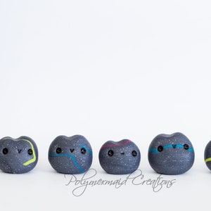 May include: Five small, gray, speckled, heart-shaped figurines with painted details. Each figurine has a different colored line design and black eyes. The figurines are arranged in a row on a white surface.