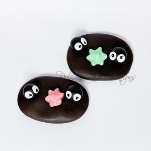 May include: Two dark brown, oval-shaped polymer clay creations with cartoon-like faces. Each has two large, white-eyed, black-pupiled eyes and a star-shaped accent in either light green or pink. The text "Polymermaid Creations" is visible.