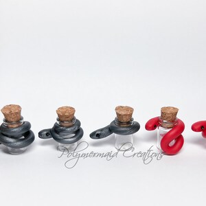 May include: Five small glass bottles with cork tops, each containing a coiled polymer clay snake. Two bottles contain gray snakes, and three bottles contain red snakes.