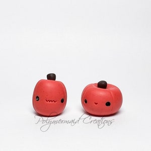 May include: Two small red clay pumpkins with black eyes and mouths. The pumpkin on the left has a slightly furrowed brow, while the pumpkin on the right has a happy expression.