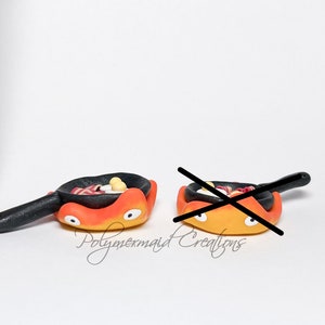 May include: Two miniature ceramic frying pans with smiling faces. The pans are orange and yellow with black handles. The pans are filled with miniature food, including bacon, eggs, and sausage.