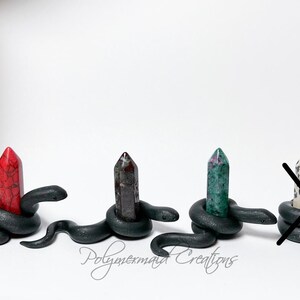May include: Four black snake sculptures, each with a different colored crystal point on top. The crystals are red, purple, green, and white with black spots. The snakes are coiled around the crystals.