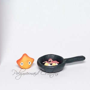 May include: A miniature orange and yellow star-shaped character with eyes and a black frying pan with a handle. The pan contains two yellow eggs and three strips of red bacon.