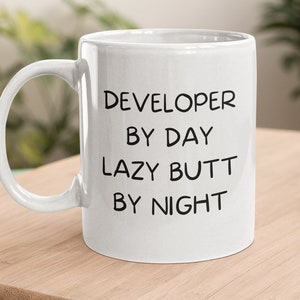 May include: White ceramic coffee mug with black text that reads "DEVELOPER BY DAY LAZY BUTT BY NIGHT".