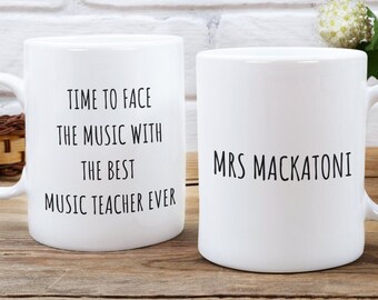 Może przedstawiać: Dwa białe ceramiczne kubki z czarnym tekstem. Jeden kubek ma napis "Time to face the music with the best music teacher ever". Drugi kubek ma napis "Mrs Mackatoni".