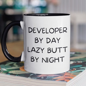 May include: White ceramic mug with a black handle and black rim. The mug has black text that reads "DEVELOPER BY DAY LAZY BUTT BY NIGHT". The mug is sitting on a book with a floral pattern.