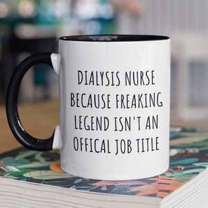 May include: A white ceramic mug with a black handle and rim. The mug features the text "DIALYSIS NURSE BECAUSE FREAKING LEGEND ISN'T AN OFFICAL JOB TITLE" in black lettering. The mug is sitting on a stack of books.