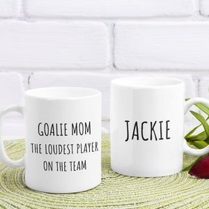 May include: Two white ceramic mugs with black text. The mug on the left says "Goalie Mom The Loudest Player On The Team". The mug on the right says "Jackie". The mugs are sitting on a green placemat with a red lily flower.