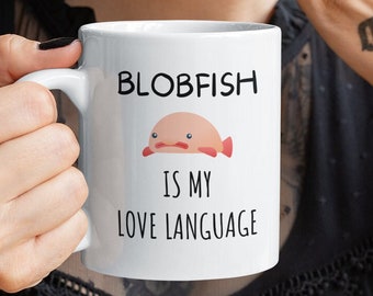 May include: White ceramic mug with a black handle. The mug features a cartoon blobfish with a pink mouth and a black moustache. The text on the mug reads "BLOBFISH IS MY LOVE LANGUAGE".