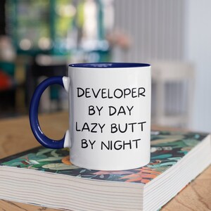 May include: White ceramic mug with a blue rim and handle. The mug has black text that reads "DEVELOPER BY DAY LAZY BUTT BY NIGHT".