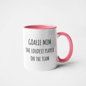 May include: White ceramic coffee mug with a pink rim and handle. The mug has black text that reads "Goalie Mom The Loudest Player On The Team."