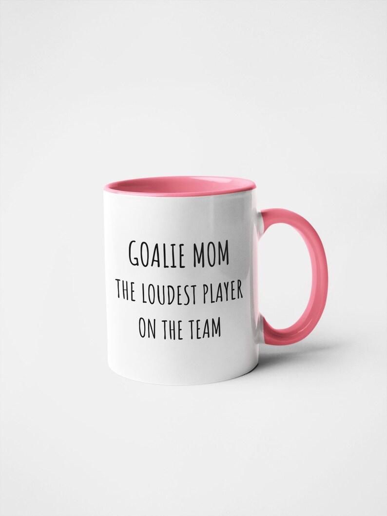 May include: White ceramic coffee mug with a pink rim and handle. The mug has black text that reads "Goalie Mom The Loudest Player On The Team."