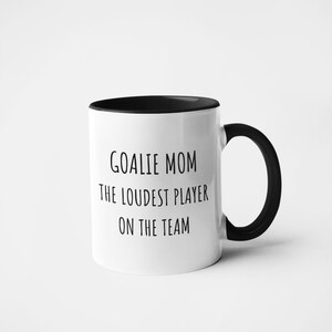 May include: White ceramic coffee mug with a black rim and handle. The mug has black text that reads "Goalie Mom The Loudest Player On The Team."