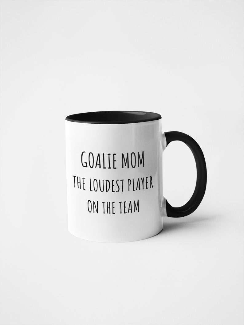 May include: White ceramic coffee mug with a black rim and handle. The mug has black text that reads "Goalie Mom The Loudest Player On The Team."