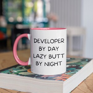 May include: White ceramic mug with a pink rim and handle. The mug has black text that reads "DEVELOPER BY DAY LAZY BUTT BY NIGHT". The mug is sitting on a book with a floral pattern.