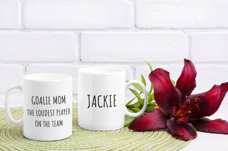 May include: Two white ceramic mugs with black text. The mug on the left says "Goalie Mom The Loudest Player On The Team". The mug on the right says "Jackie". The mugs are sitting on a green placemat with a red lily flower.