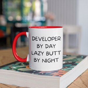May include: White ceramic mug with a red rim and handle. The mug has black text that reads "DEVELOPER BY DAY LAZY BUTT BY NIGHT". The mug is sitting on a book with a floral pattern.