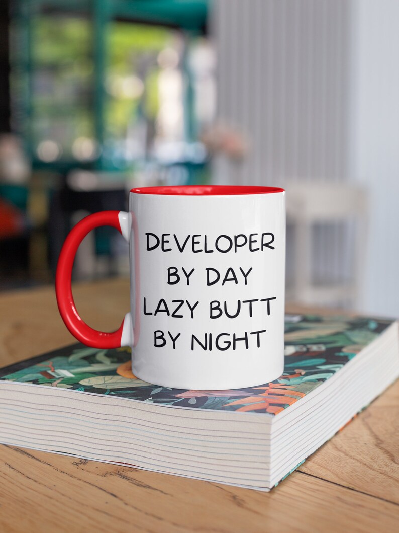 May include: White ceramic mug with a red rim and handle. The mug has black text that reads "DEVELOPER BY DAY LAZY BUTT BY NIGHT". The mug is sitting on a book with a floral pattern.