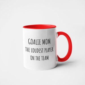 May include: White ceramic coffee mug with a red rim and handle. The mug has black text that reads "Goalie Mom The Loudest Player On The Team."