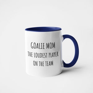 May include: White ceramic coffee mug with a blue rim and handle. The mug has black text that reads "Goalie Mom The Loudest Player On The Team."
