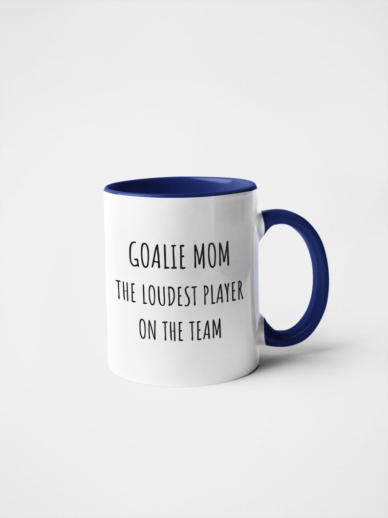 May include: White ceramic coffee mug with a blue rim and handle. The mug has black text that reads "Goalie Mom The Loudest Player On The Team."