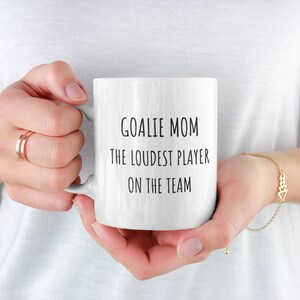 May include: White ceramic coffee mug with black text that reads "Goalie Mom The Loudest Player On The Team".