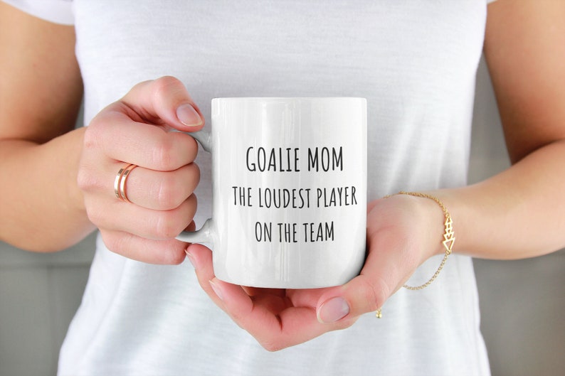 May include: White ceramic coffee mug with black text that reads "Goalie Mom The Loudest Player On The Team".