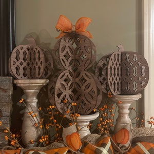 May include: Three wooden pumpkins with intricate cut-out designs, decorated with an orange bow. The pumpkins are displayed on white pedestals with a brown and orange plaid fabric and small orange pumpkins in the foreground.