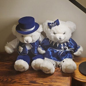 Puede incluir: Dos osos de peluche blancos vestidos con ropa formal azul. El oso macho lleva un sombrero de copa azul con una banda plateada y un chaleco azul con ribetes plateados. La osa hembra lleva un vestido azul con ribetes plateados y un ribete de encaje blanco. Ambos osos tienen pelaje blanco y ojos negros.