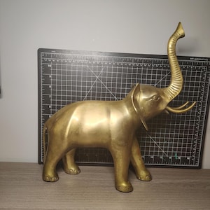 May include: A gold-coloured brass elephant ornament with its trunk raised. The elephant has tusks and is standing on four legs. The ornament is on a wooden surface with a black cutting mat in the background.