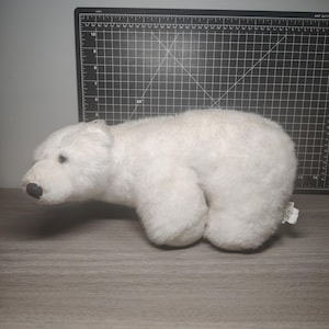 以下が含まれることがあります： 白いぬいぐるみホッキョクグマ。クマは黒い鼻と小さな黒い目をしています。おもちゃは柔らかくふわふわした素材でできており、木製の表面に置かれています。背景には黒いカッティングマットがあります。