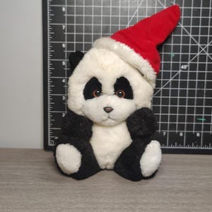 Può includere: Un orsacchiotto panda con un cappello da Babbo Natale rosso. Il panda ha la pelliccia bianca e nera, con macchie nere intorno agli occhi e sulle orecchie, braccia e gambe. L'orso è seduto in posizione eretta, con le braccia appoggiate.